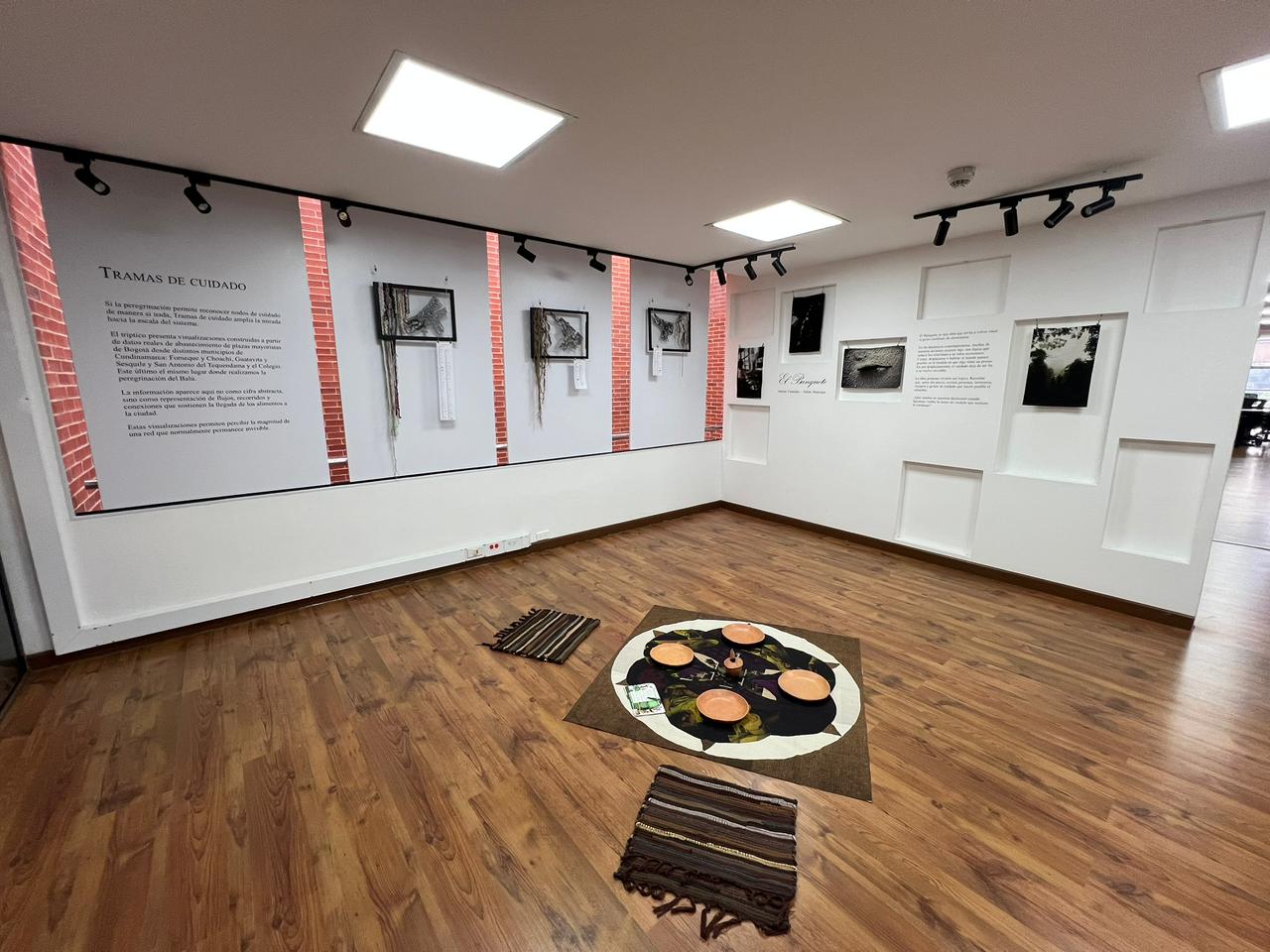 Vista general de la sala: el tríptico y el texto de sala en la pared izquierda, fotografías enmarcadas al fondo y el mandala con cuencos de barro en el centro del piso.