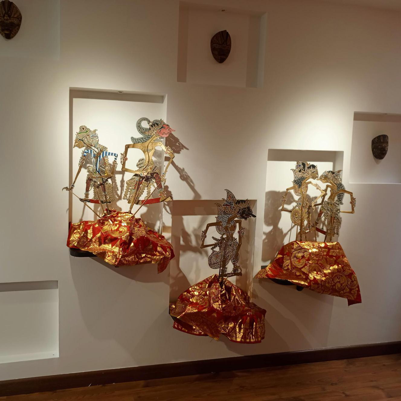 Figuras de wayang kulit en nichos iluminados del muro, sobre textiles rojos y dorados.