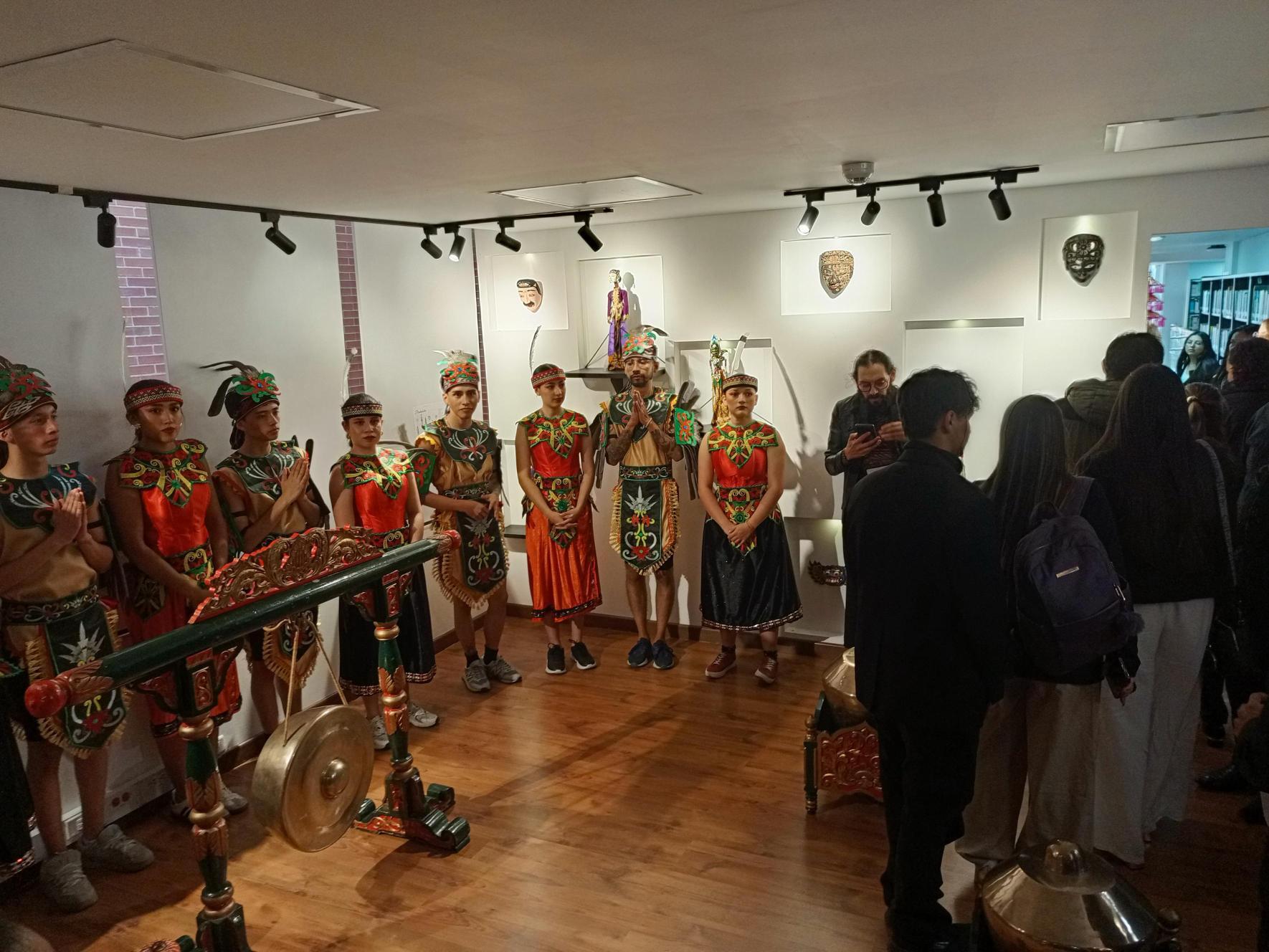 Grupo con trajes tradicionales de pie en la sala, con el gong en primer plano y el público a la derecha.